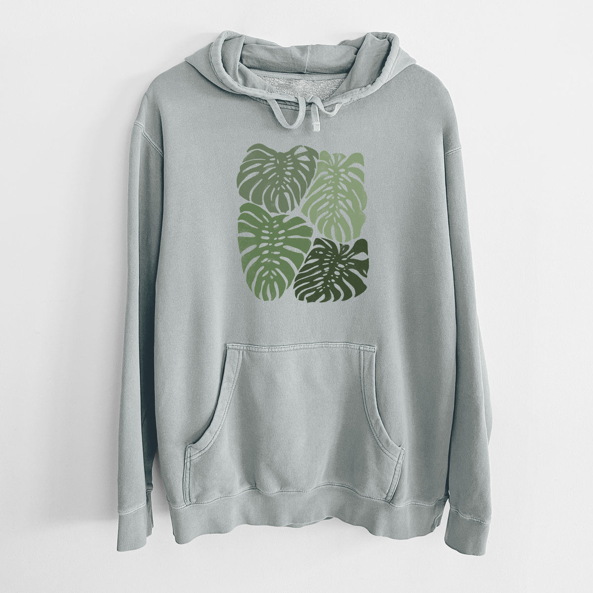 Sage green pigment dyed hoodie laid flat, printed with a full-color illustration of large green Monstera leaves filling a square.