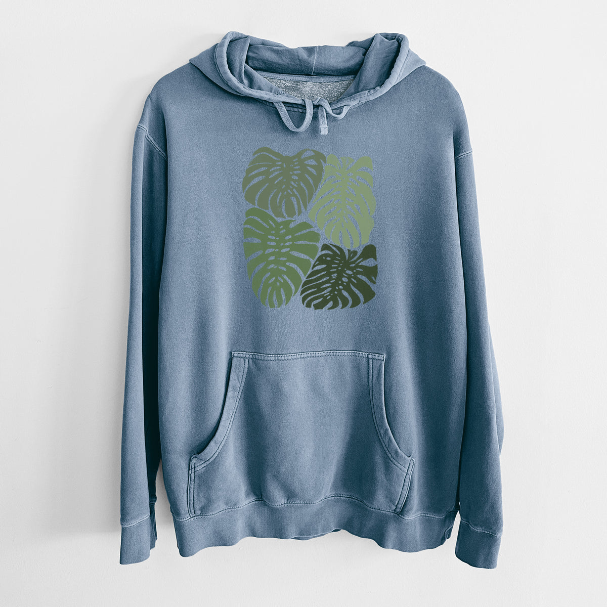 Slate blue pigment dyed hoodie laid flat, printed with a full-color illustration of large green Monstera leaves filling a square.