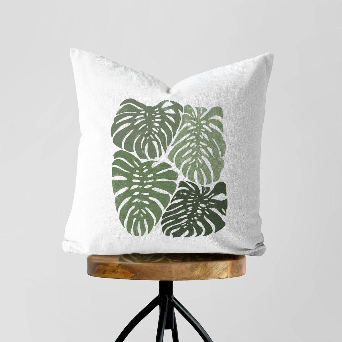 White pillow cover printed with a full-color illustration of large green Monstera leaves filling a square, on a wooden stool.