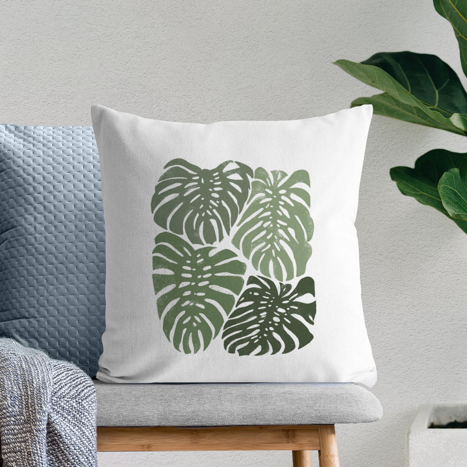 White pillow cover printed with a full-color illustration of large green Monstera leaves filling a square, on a bench beside plants.