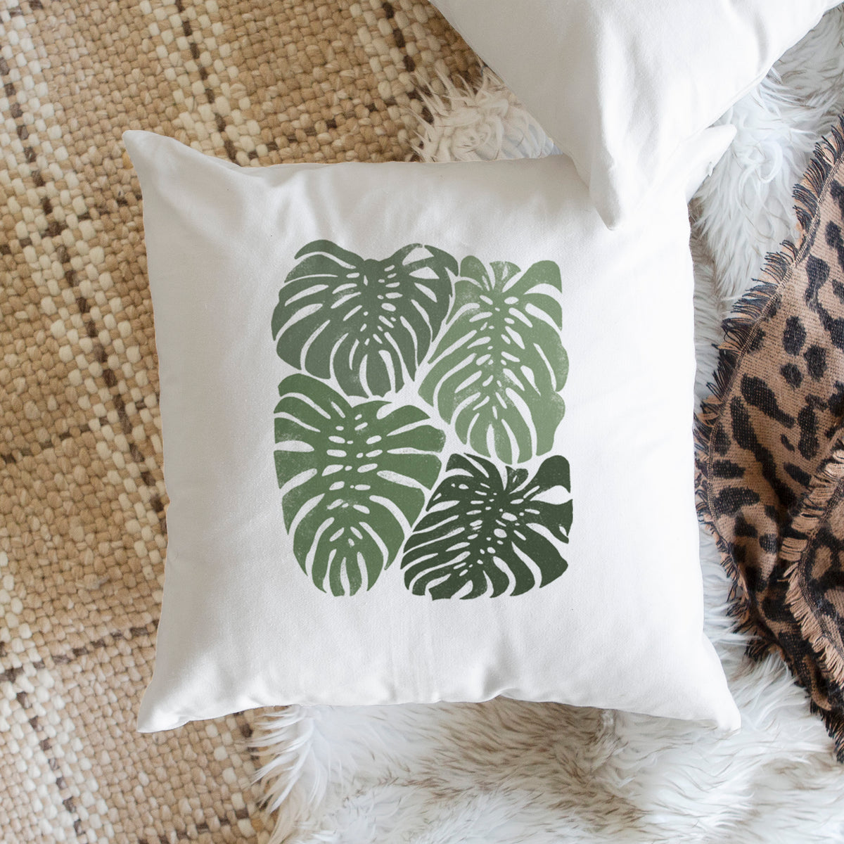 White pillow cover printed with a full-color illustration of large green Monstera leaves filling a square, on a woven chair.