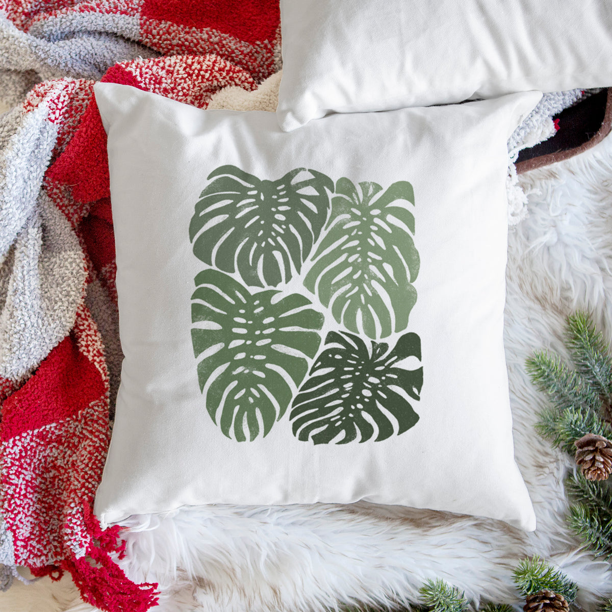 White pillow cover printed with a full-color illustration of large green Monstera leaves filling a square, on a woven chair.