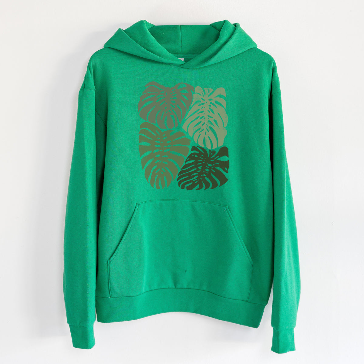 Monstera Vibes - Statement Midweight Hoodie in Kelly Green, hanging on a white wall, with a front kangaroo pocket and no drawcords.