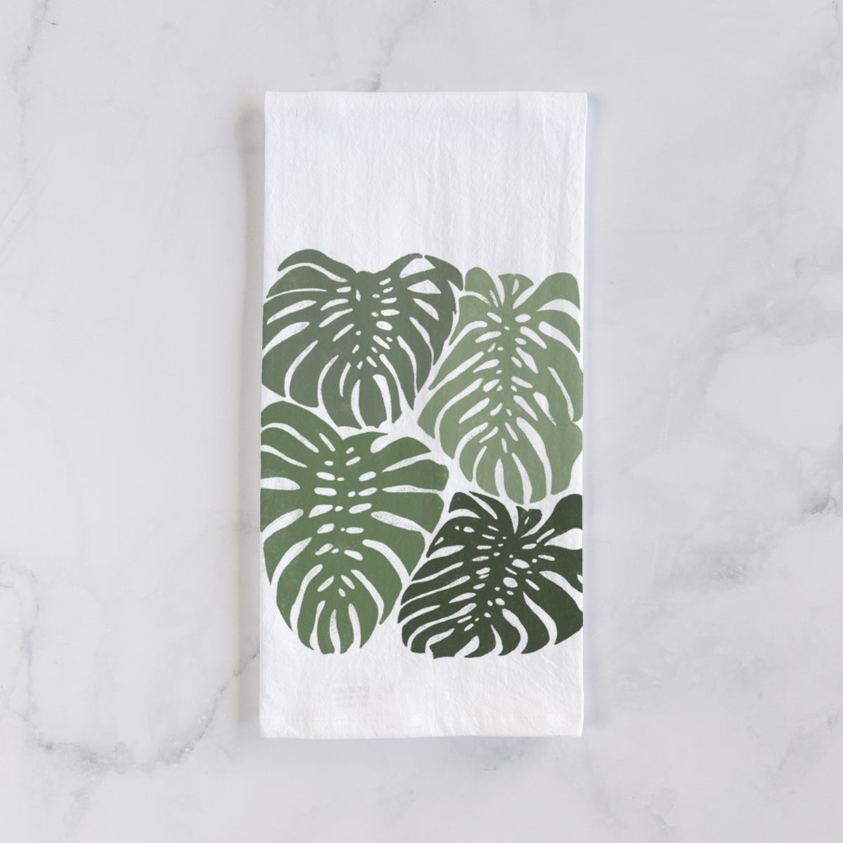 White cotton tea towel laid flat on marble, printed with a full-color illustration of large green Monstera leaves filling a square.