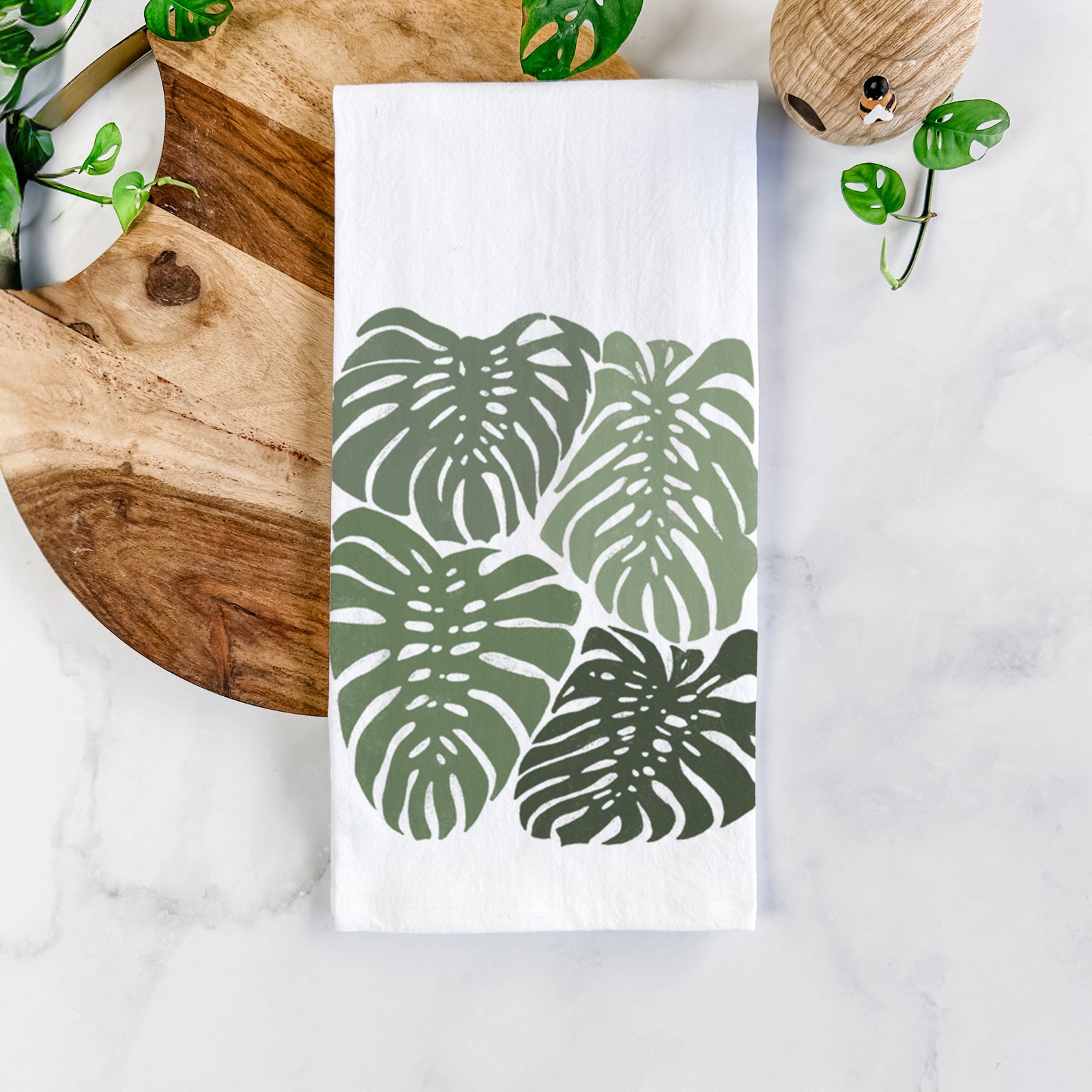 White cotton tea towel printed with a full-color illustration of large green Monstera leaves filling a square, on a marble counter beside a round wooden cutting board and greenery.