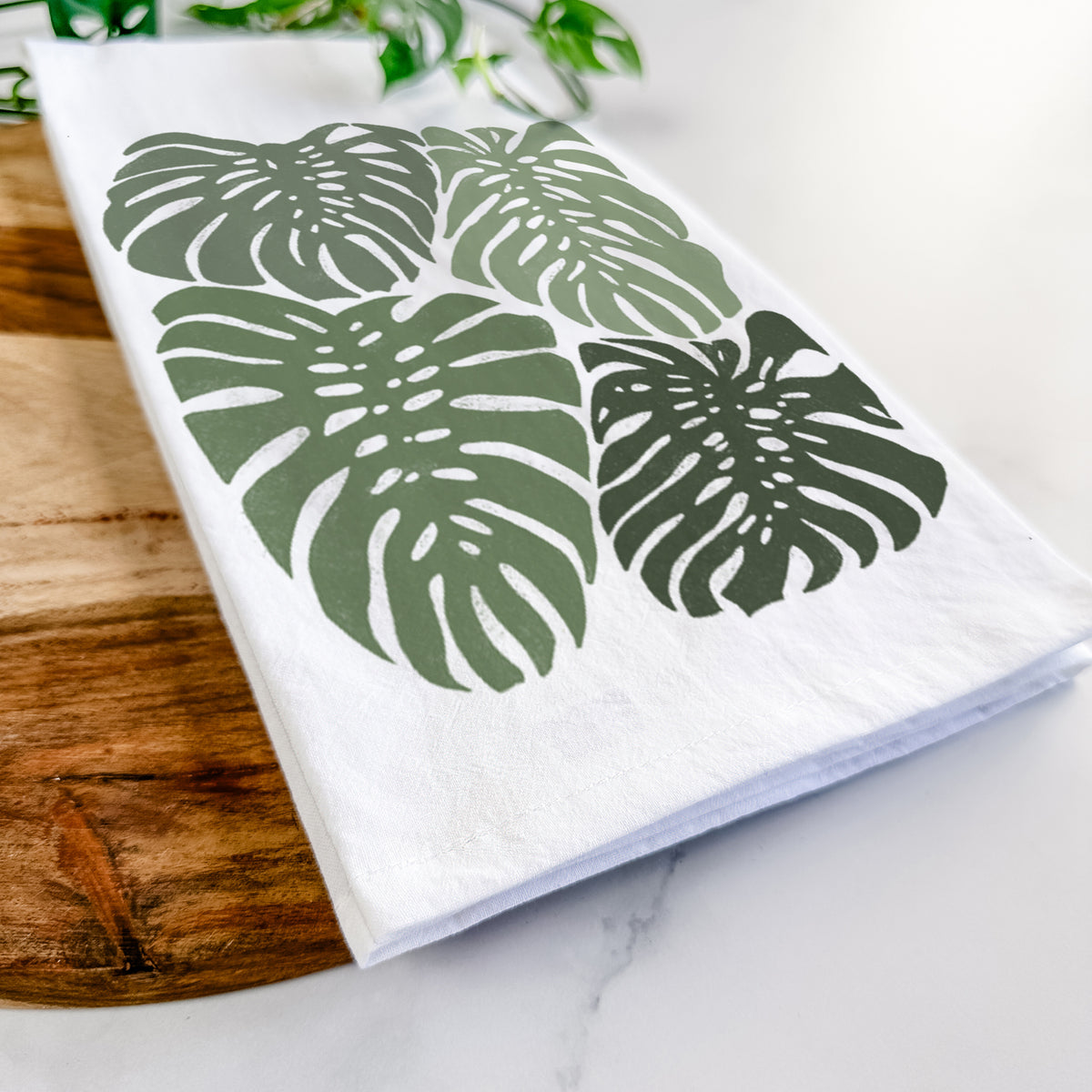 Close-up of a folded white tea towel on a wooden board, showing a full-color illustration of large green Monstera leaves filling a square.