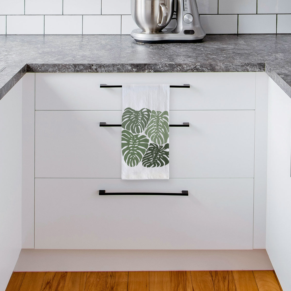 White tea towel printed with a full-color illustration of large green Monstera leaves filling a square, hanging from a drawer handle in a bright white kitchen.