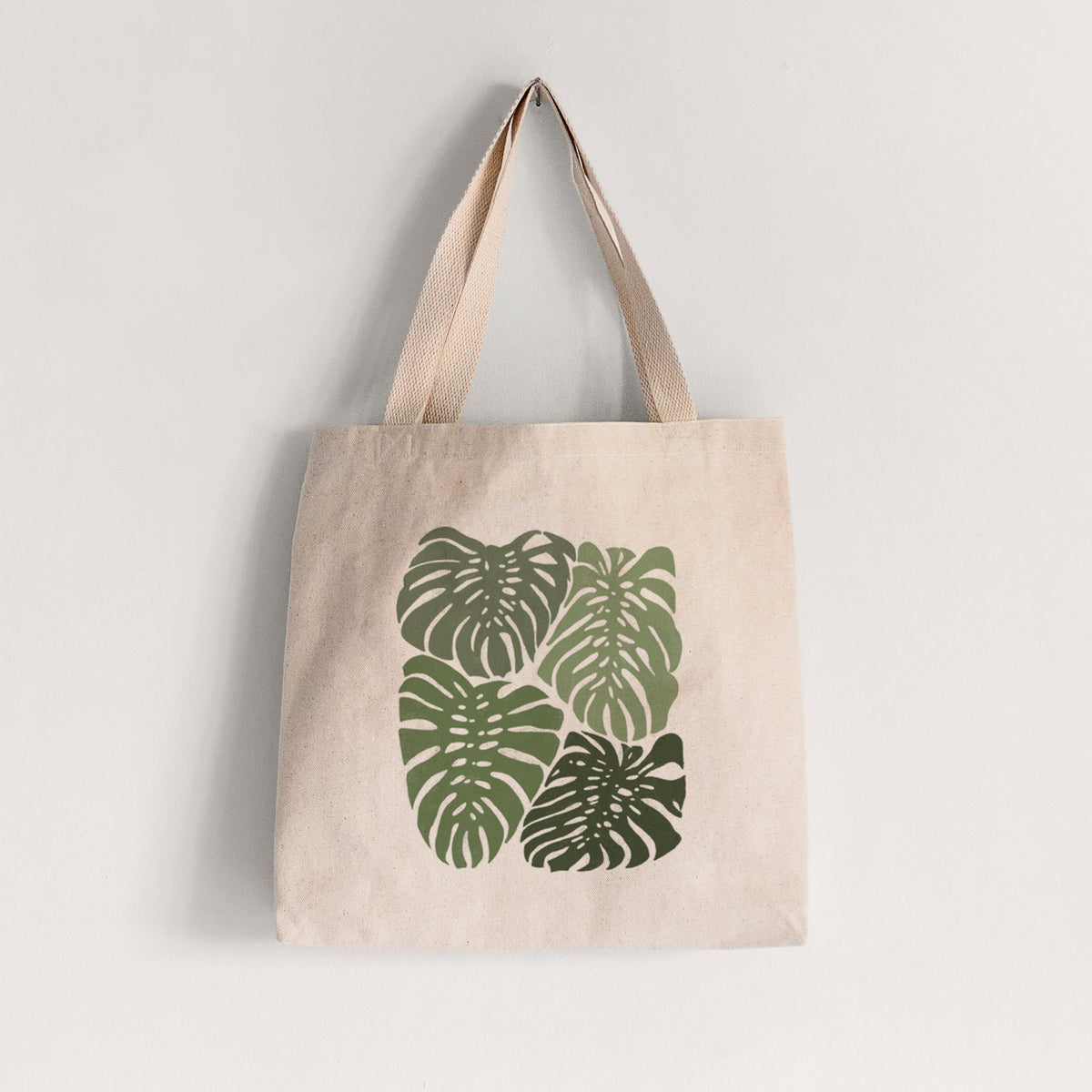 Natural canvas tote bag printed with a full-color illustration of large green Monstera leaves filling a square.