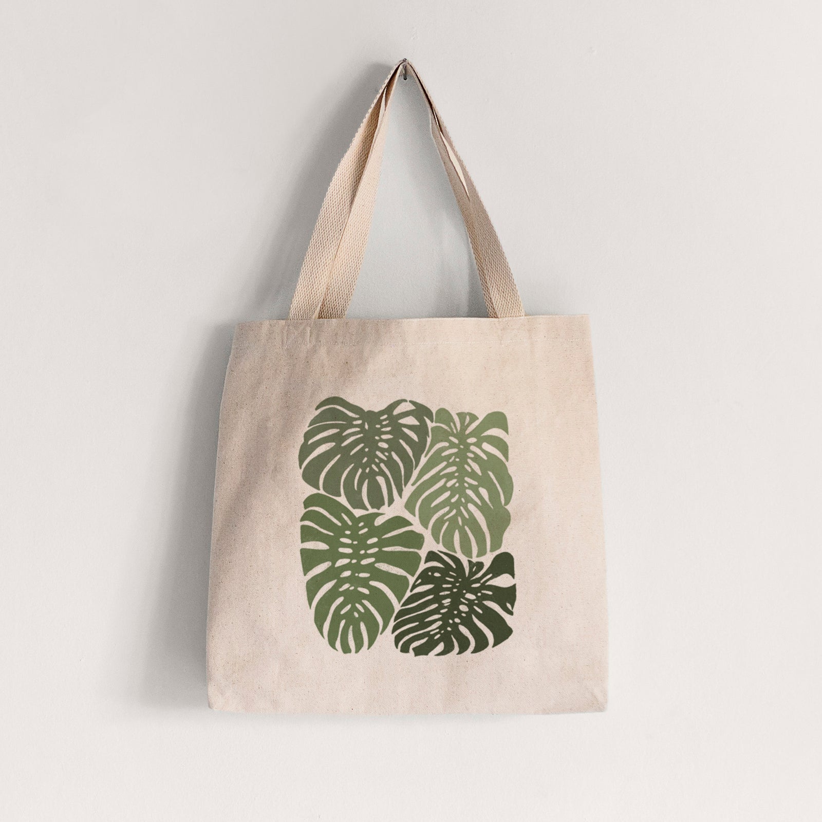 Natural canvas tote bag printed with a full-color illustration of large green Monstera leaves filling a square.