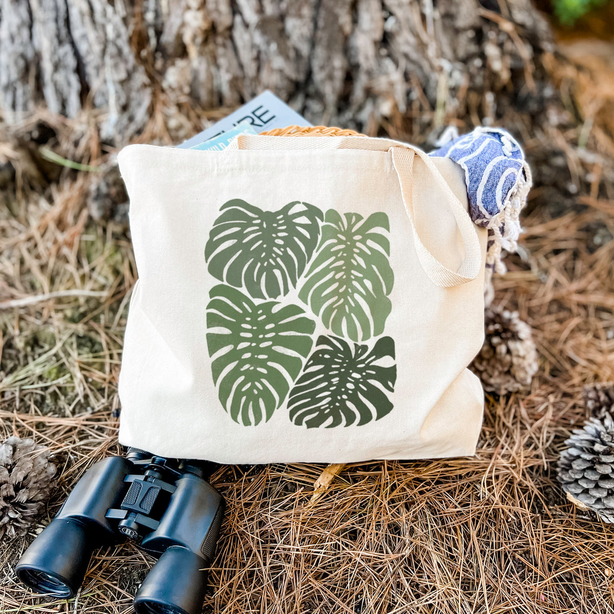 Canvas tote bag printed with a full-color illustration of large green Monstera leaves filling a square, resting on dry grass beside a pair of binoculars.