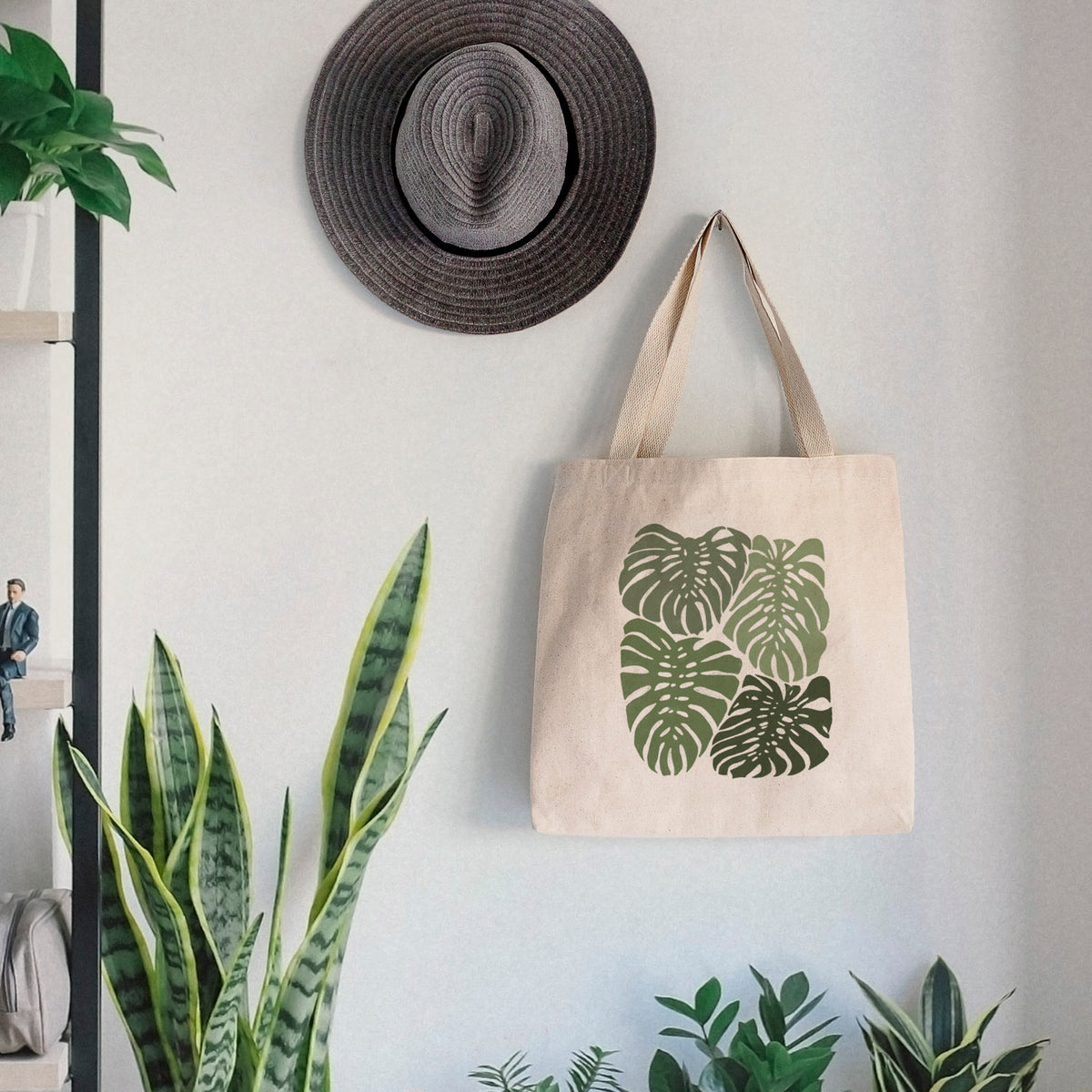 Canvas tote bag printed with a full-color illustration of large green Monstera leaves filling a square, hanging on a wall hook below a hat, next to potted plants.