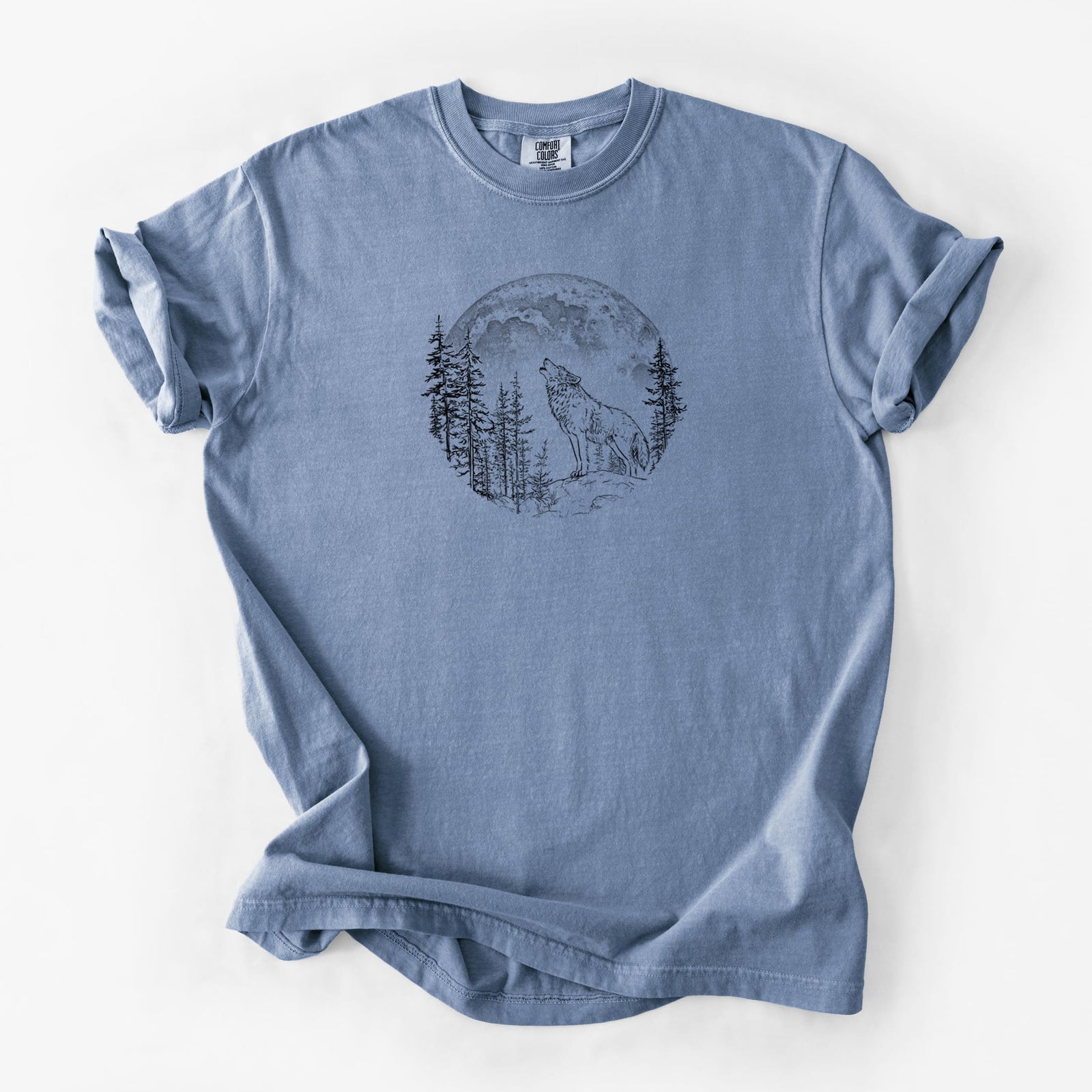 Blue jean Comfort Colors t-shirt laid flat, printed with a black ink wolf howling in front of a full moon, surrounded by pine trees.