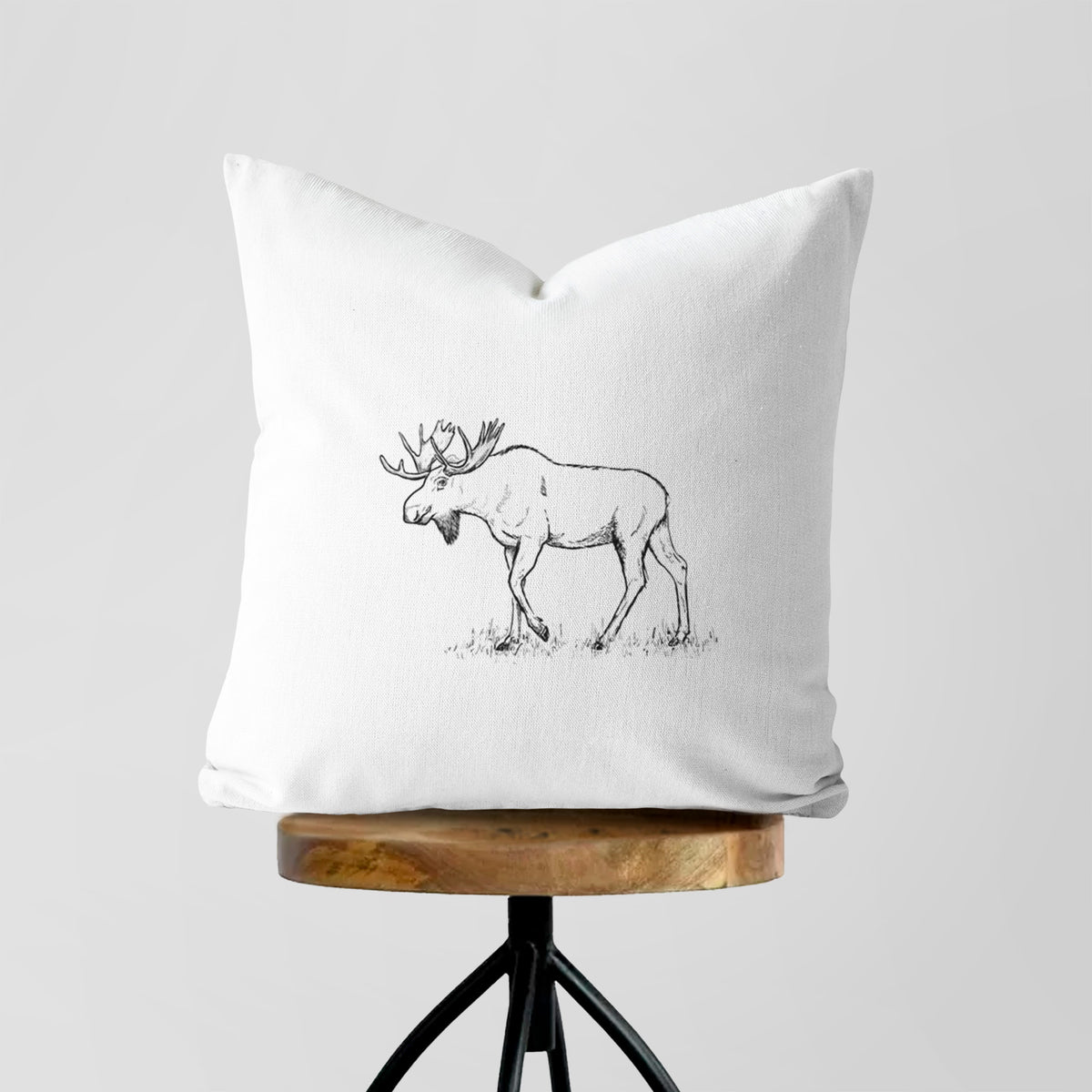 White pillow cover printed with a black ink illustration of a bull moose with broad antlers walking, on a wooden stool.