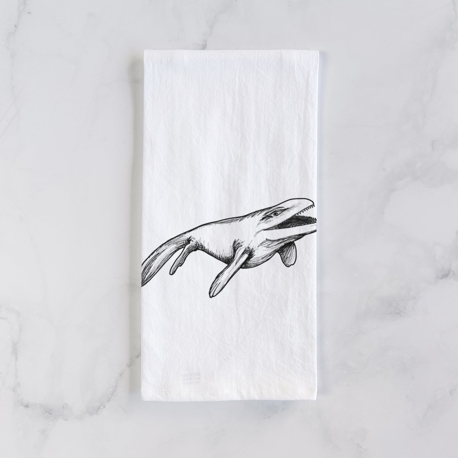 White cotton tea towel printed with a black ink illustration of a Mosasaur swimming, on a marble counter beside a round wooden cutting board and greenery.