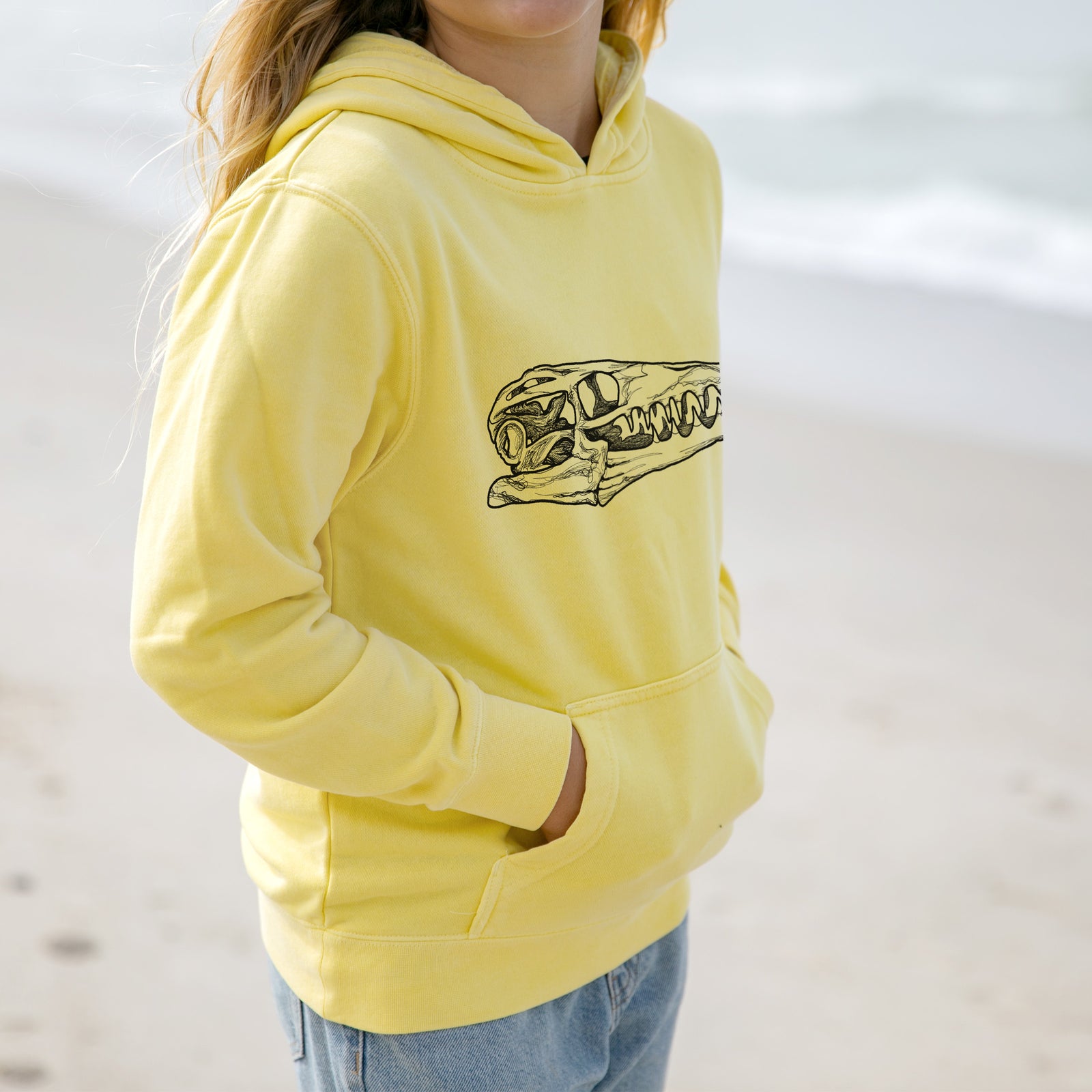 Boy wearing a youth pigment dyed hoodie printed with a detailed black ink illustration of a Mosasaur skull.