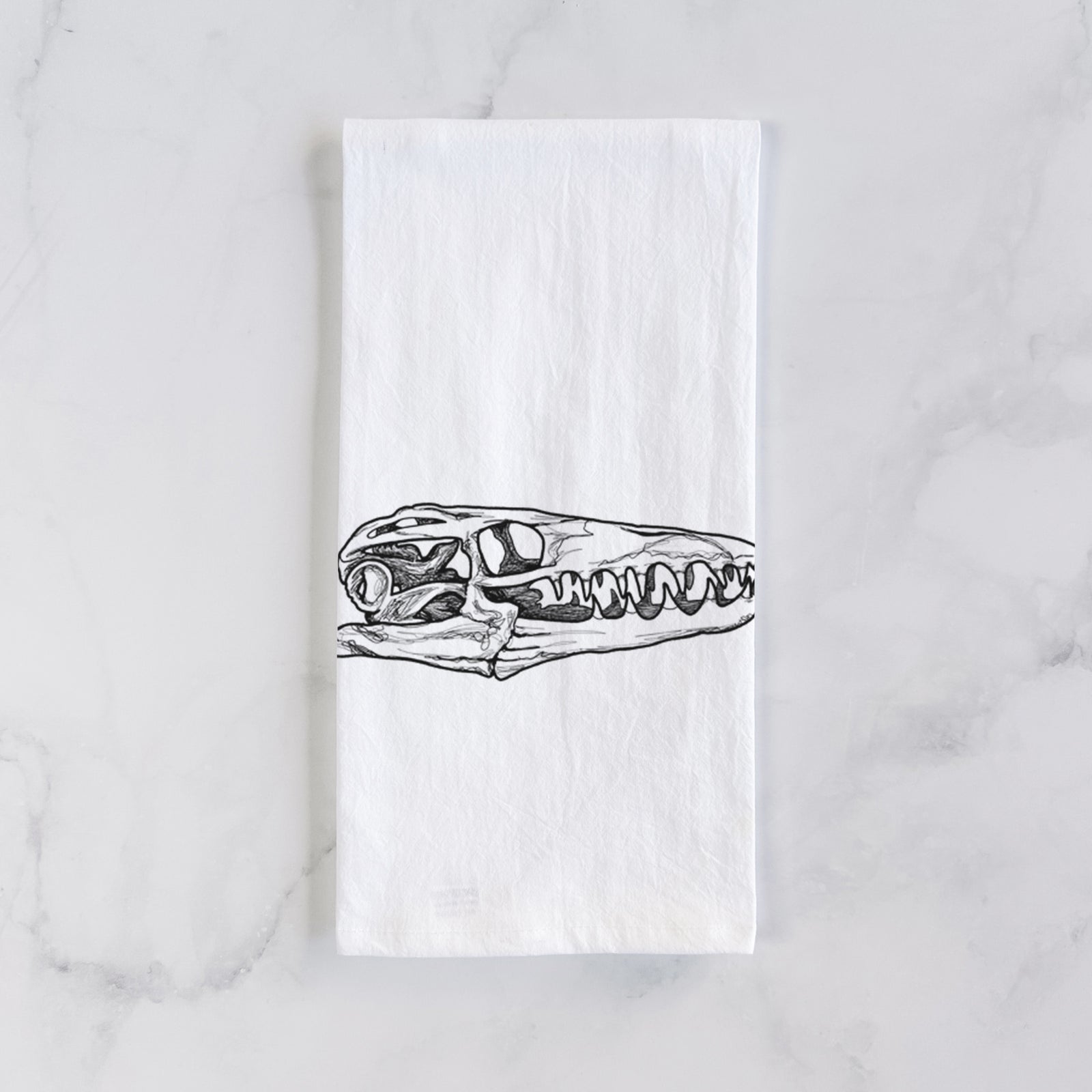 White cotton tea towel printed with a detailed black ink illustration of a Mosasaur skull, on a marble counter beside a round wooden cutting board and greenery.