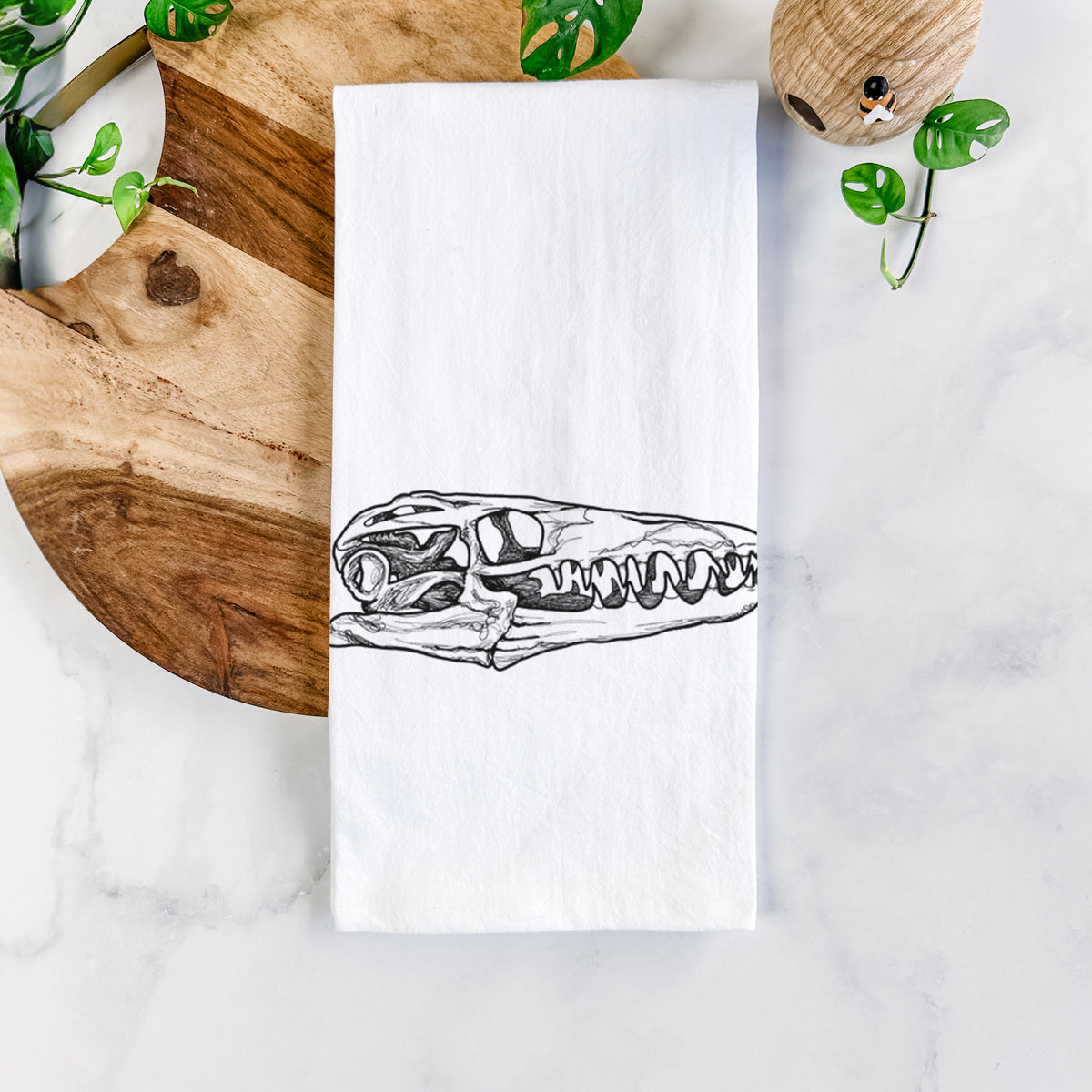 White cotton tea towel printed with a detailed black ink illustration of a Mosasaur skull, on a marble counter beside a round wooden cutting board and greenery.