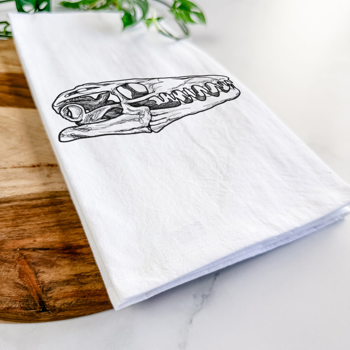Close-up of a folded white tea towel on a wooden board, showing a detailed black ink illustration of a Mosasaur skull.