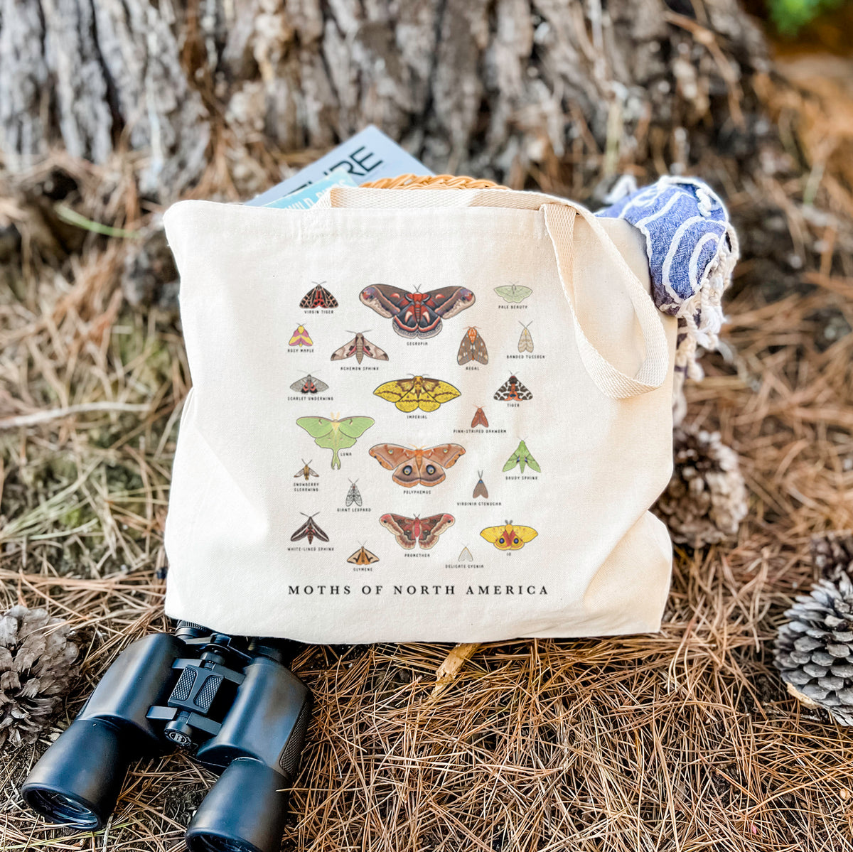 Canvas tote bag printed with a full-color chart of North American moths, each labeled with its species, resting on dry grass beside a pair of binoculars.