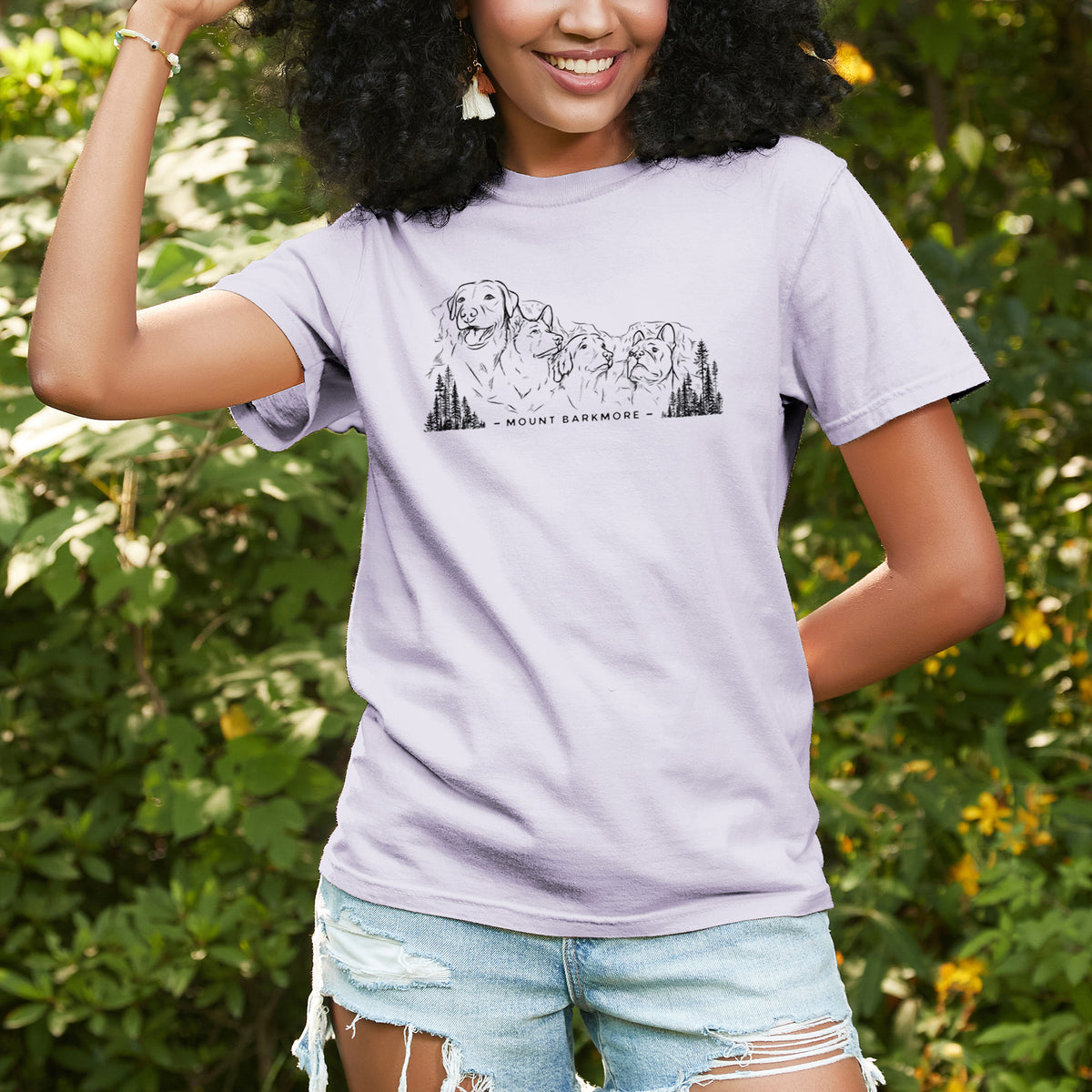 Woman wearing a Comfort Colors t-shirt printed with a black ink illustration of four dog faces carved into a mountain, with the words Mount Barkmore, outdoors.