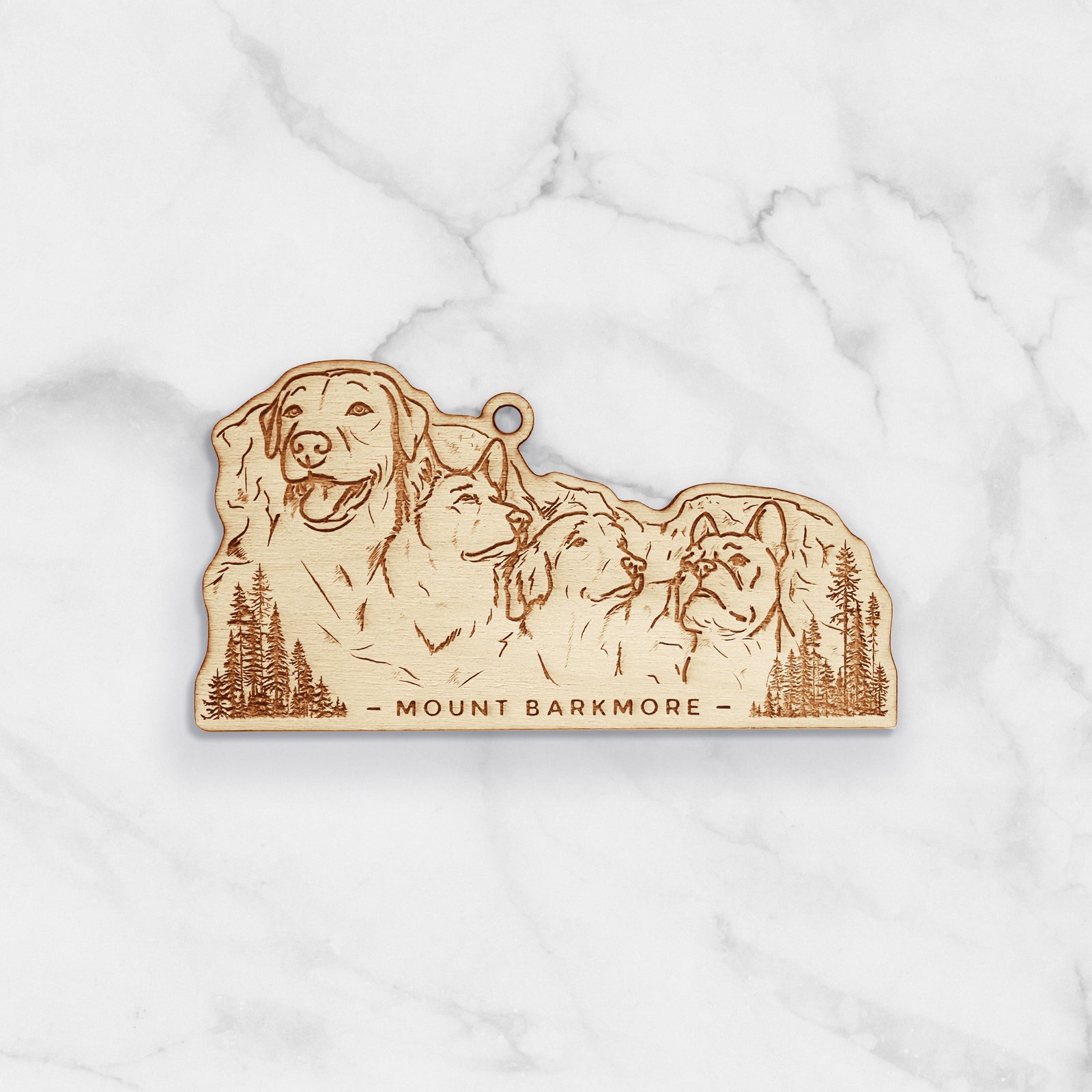 Wooden ornament featuring a engraving of four dog faces carved into a mountain, with the words Mount Barkmore, on a knit blanket with pine cones.