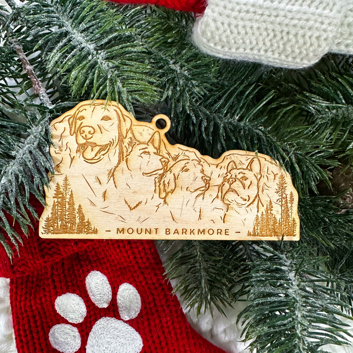 Wooden ornament featuring a engraving of four dog faces carved into a mountain, with the words Mount Barkmore, on a knit blanket with pine cones.