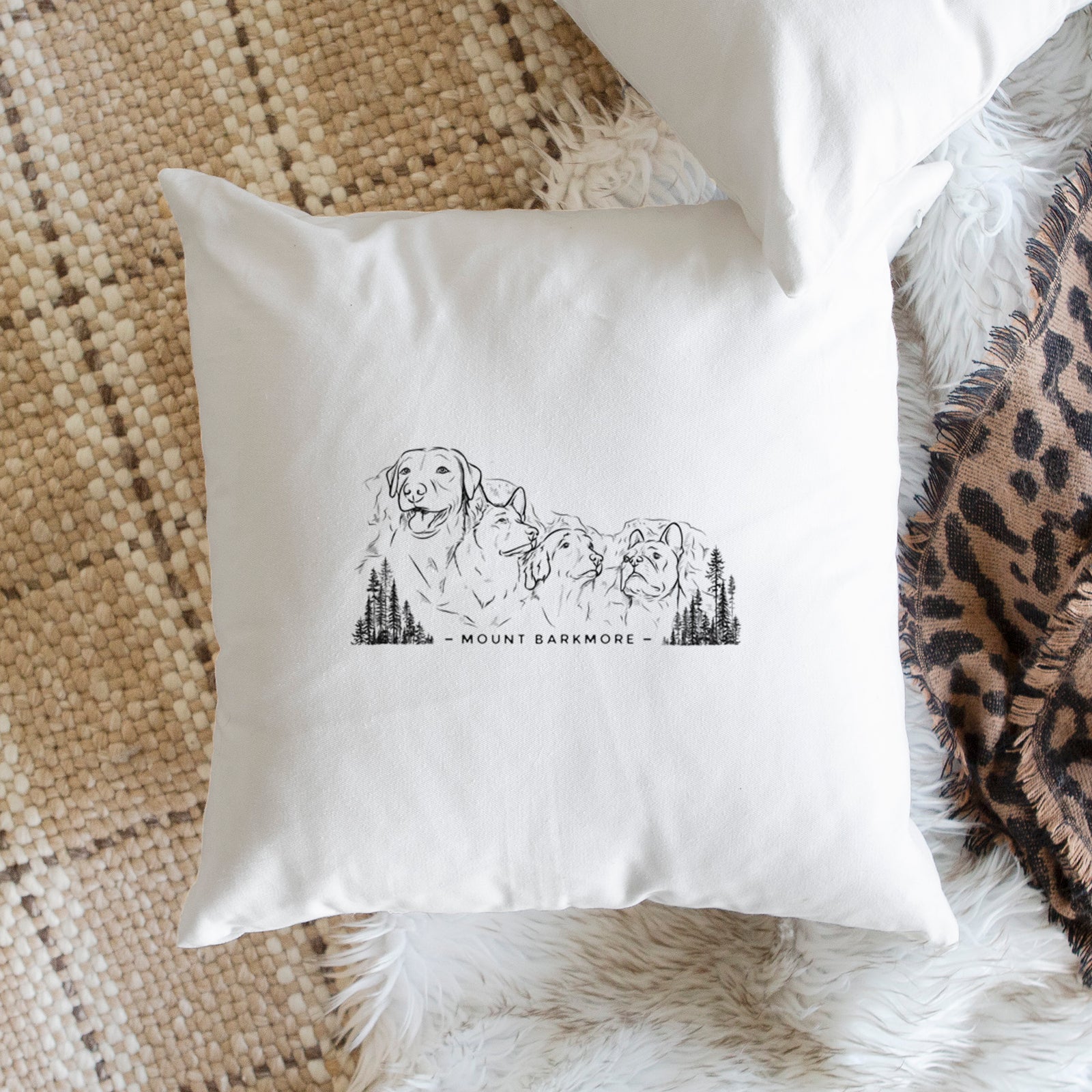 White pillow cover printed with a black ink illustration of four dog faces carved into a mountain, with the words Mount Barkmore, on a bench beside plants.