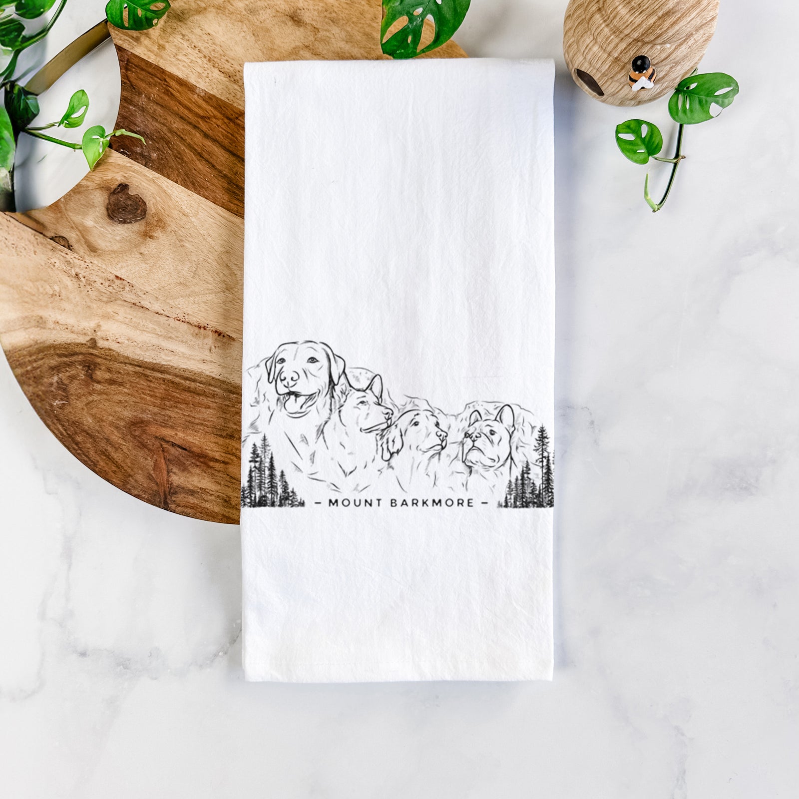White cotton tea towel printed with a black ink illustration of four dog faces carved into a mountain, with the words Mount Barkmore, on a marble counter beside a round wooden cutting board and greenery.