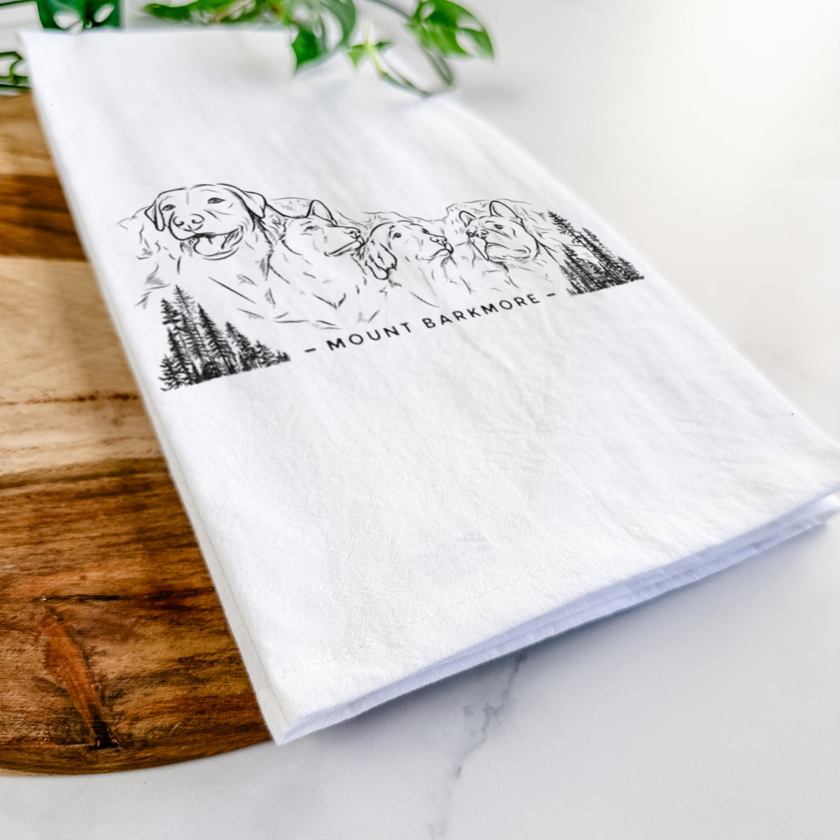 Close-up of a folded white tea towel on a wooden board, showing a black ink illustration of four dog faces carved into a mountain, with the words Mount Barkmore.