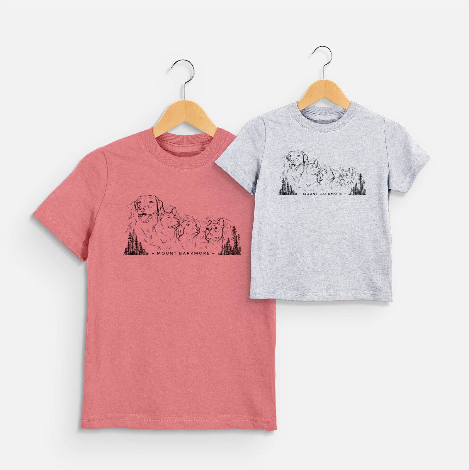 Two youth t-shirts in different colors on hangers, printed with a black ink illustration of four dog faces carved into a mountain, with the words Mount Barkmore.
