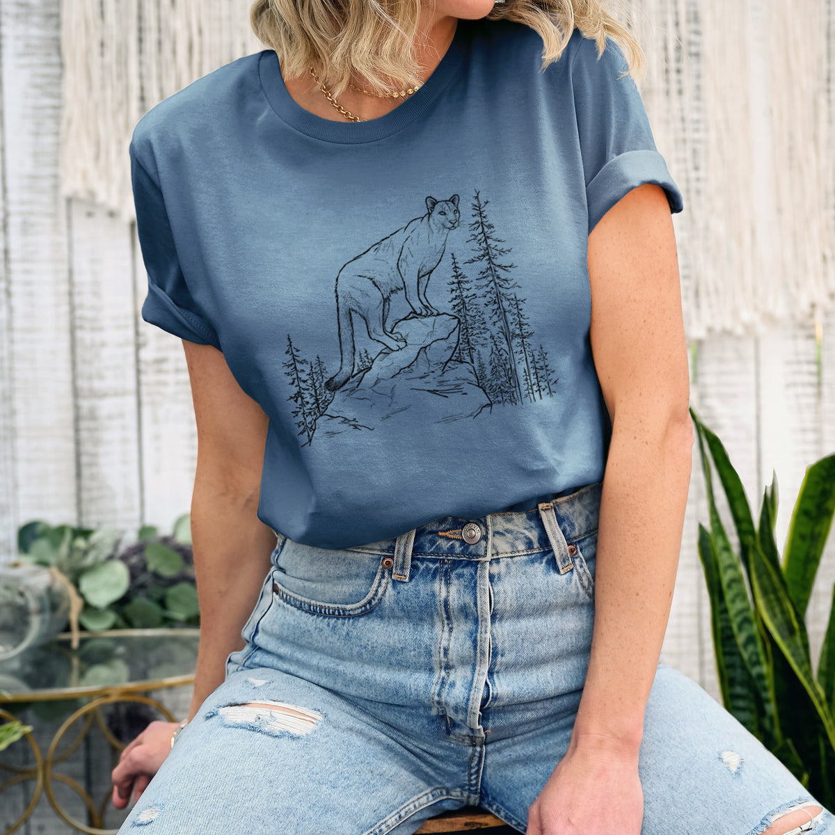 Woman wearing a steel blue lightweight cotton t-shirt printed with a black ink illustration of a mountain lion standing on a rocky ledge among pine trees.