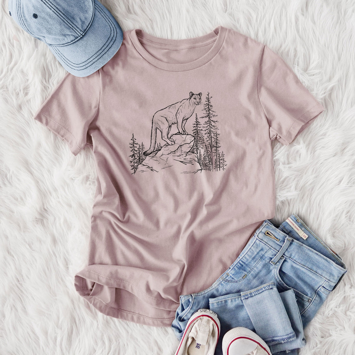 Women's relaxed fit cotton t-shirt printed with a black ink illustration of a mountain lion standing on a rocky ledge among pine trees, styled flat on a white faux fur rug with jeans.
