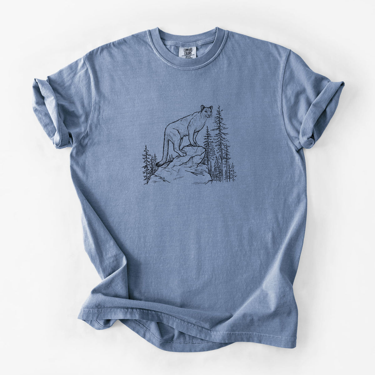 Blue jean Comfort Colors t-shirt laid flat, printed with a black ink illustration of a mountain lion standing on a rocky ledge among pine trees.