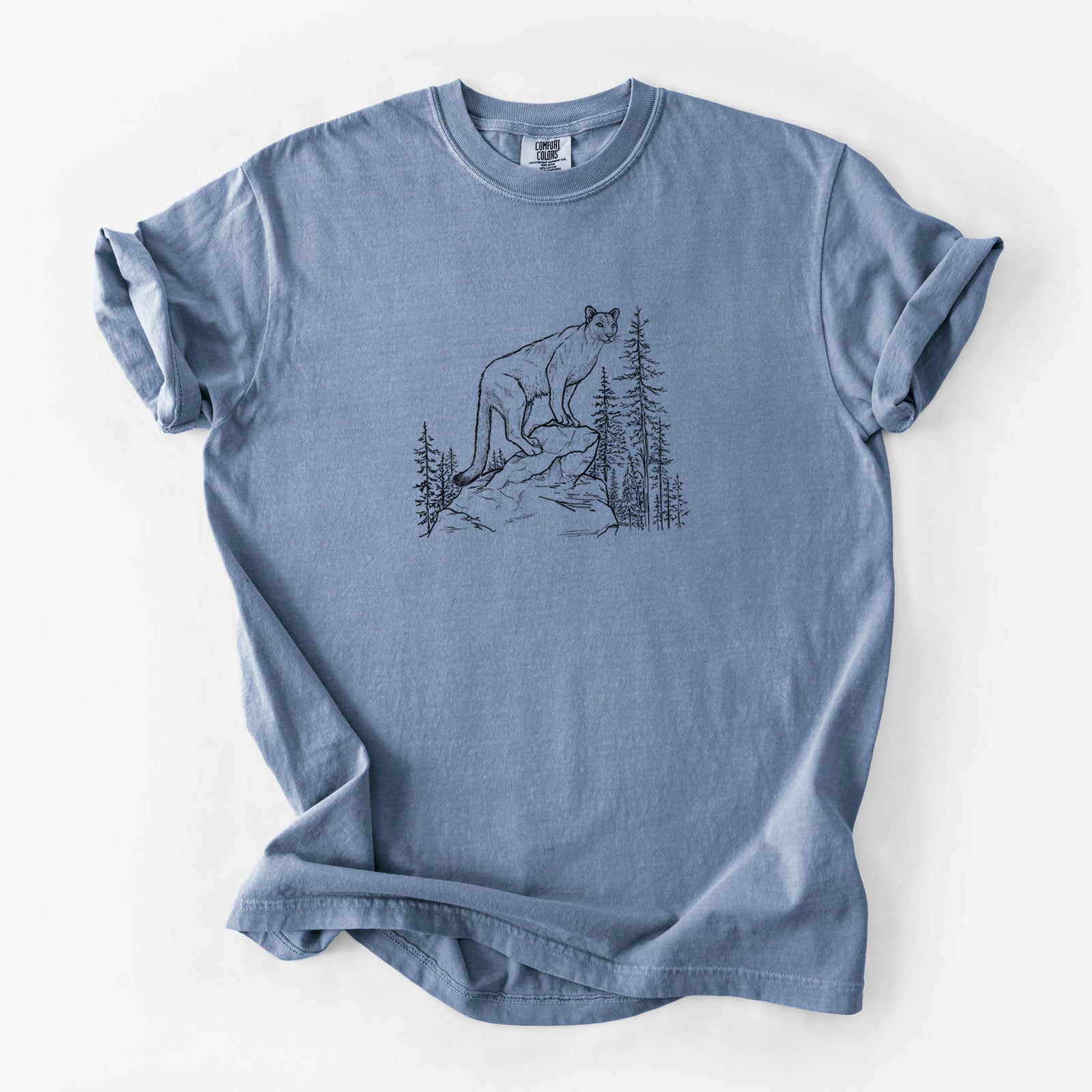 Blue jean Comfort Colors t-shirt laid flat, printed with a black ink illustration of a mountain lion standing on a rocky ledge among pine trees.
