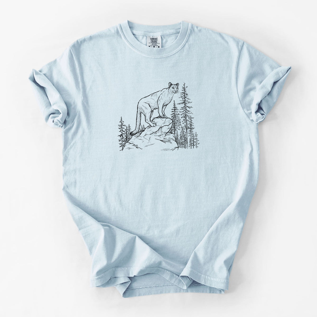 Chambray blue Comfort Colors t-shirt laid flat, printed with a black ink illustration of a mountain lion standing on a rocky ledge among pine trees.