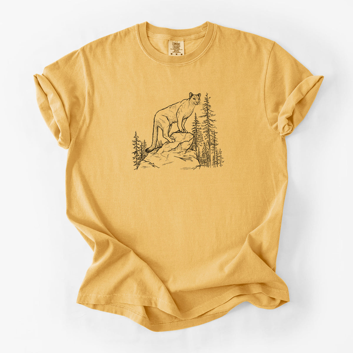 Golden wheat yellow Comfort Colors t-shirt laid flat, printed with a black ink illustration of a mountain lion standing on a rocky ledge among pine trees.