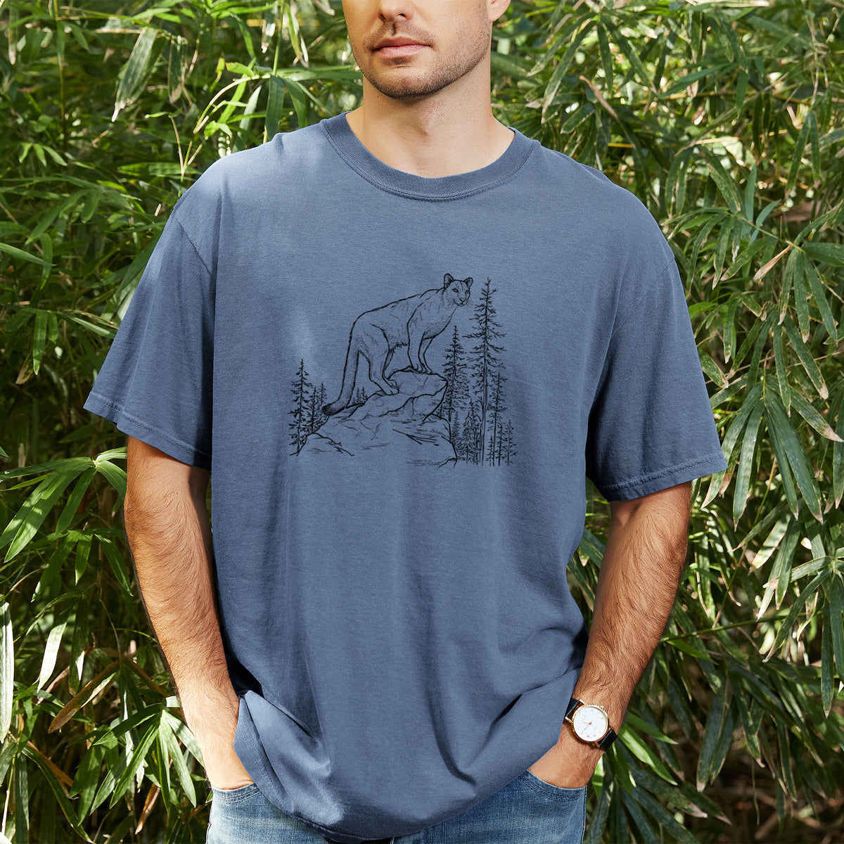 Man wearing a Comfort Colors t-shirt printed with a black ink illustration of a mountain lion standing on a rocky ledge among pine trees, outdoors.