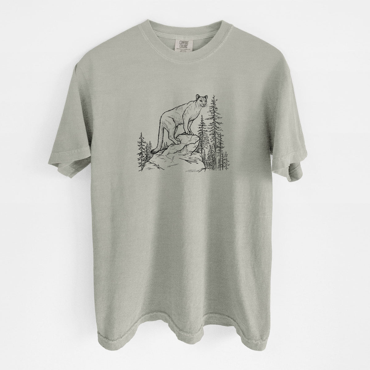 Sandstone beige Comfort Colors t-shirt laid flat, printed with a black ink illustration of a mountain lion standing on a rocky ledge among pine trees.