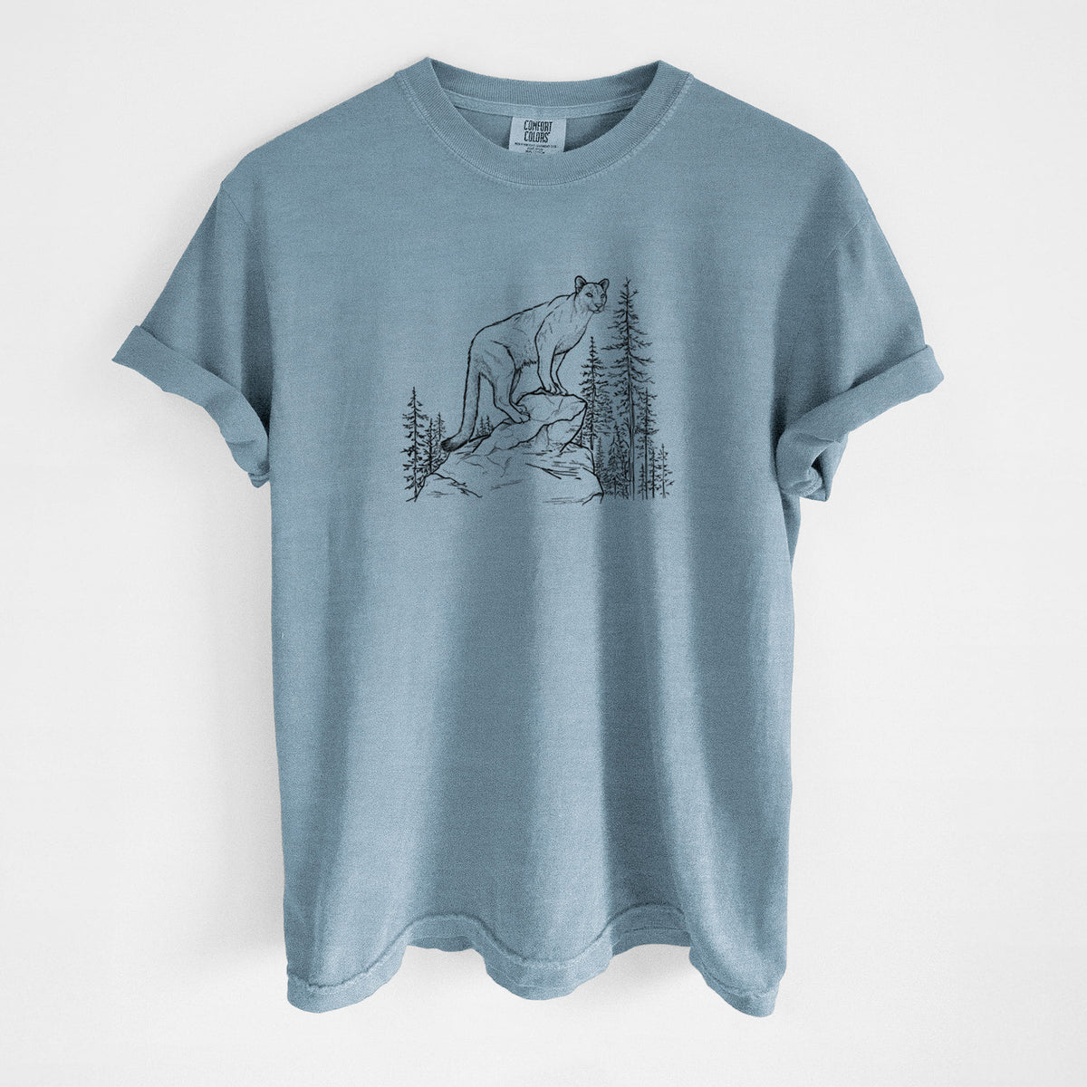 Steel blue Comfort Colors t-shirt laid flat, printed with a black ink illustration of a mountain lion standing on a rocky ledge among pine trees.