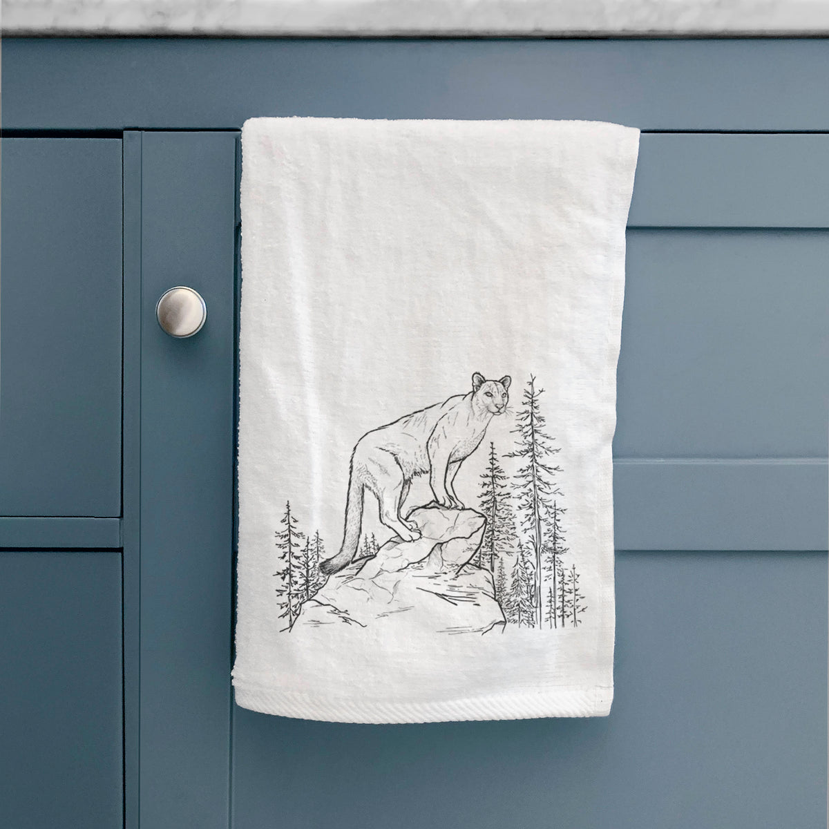 White hand towel printed with a black ink illustration of a mountain lion standing on a rocky ledge among pine trees, hanging on a cabinet door.