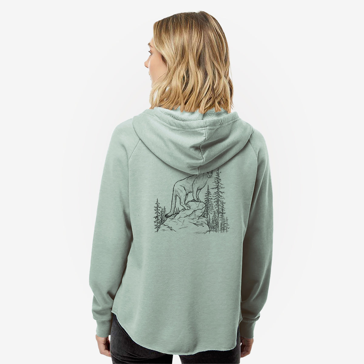 Woman wearing a sage green women's Cali Wave zip-up sweatshirt printed with a black ink illustration of a mountain lion standing on a rocky ledge among pine trees, seen from behind.