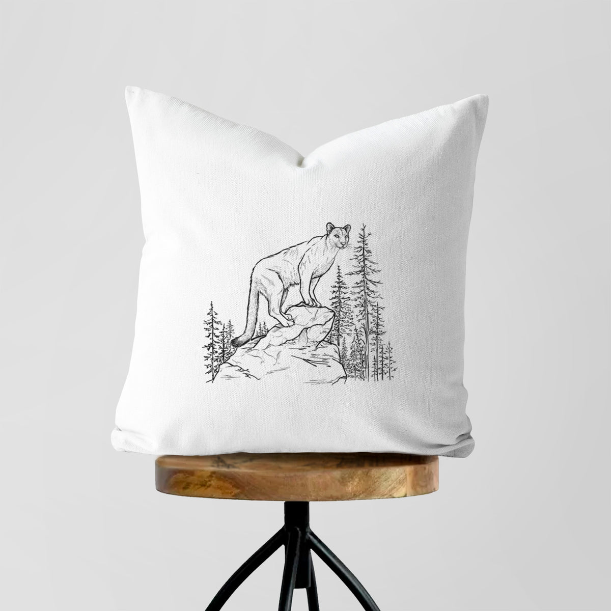 White pillow cover printed with a black ink illustration of a mountain lion standing on a rocky ledge among pine trees, on a wooden stool.