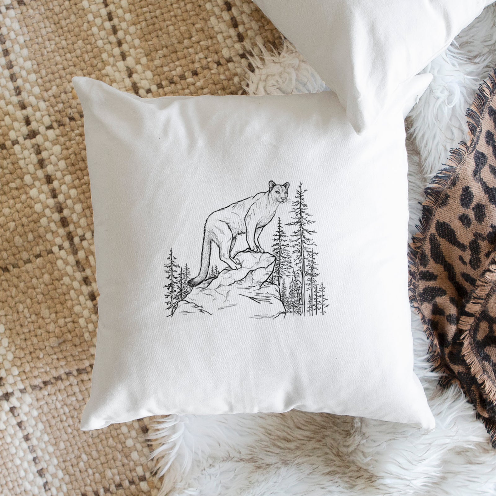 White pillow cover printed with a black ink illustration of a mountain lion standing on a rocky ledge among pine trees, on a bench beside plants.