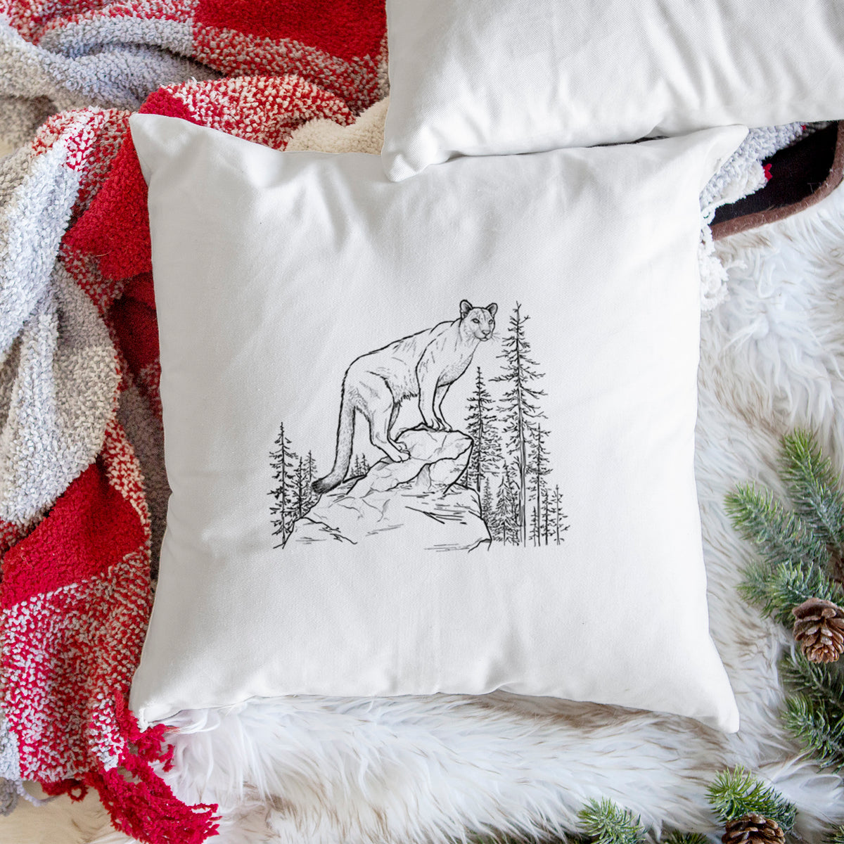 White pillow cover printed with a black ink illustration of a mountain lion standing on a rocky ledge among pine trees, on a woven chair.