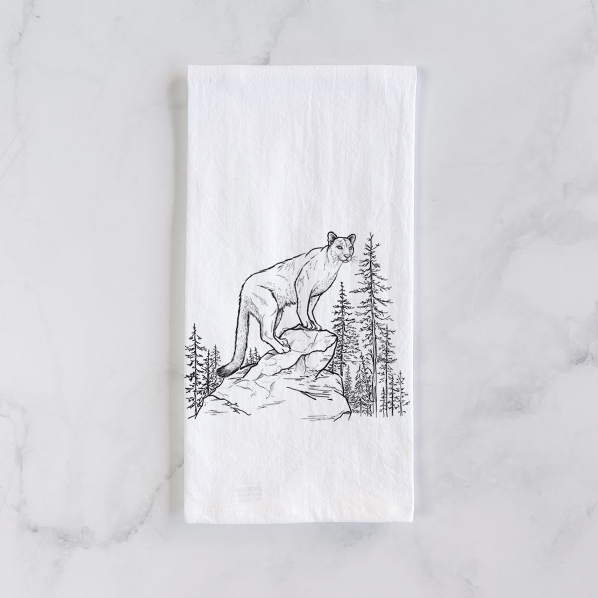 White cotton tea towel laid flat on marble, printed with a black ink illustration of a mountain lion standing on a rocky ledge among pine trees.