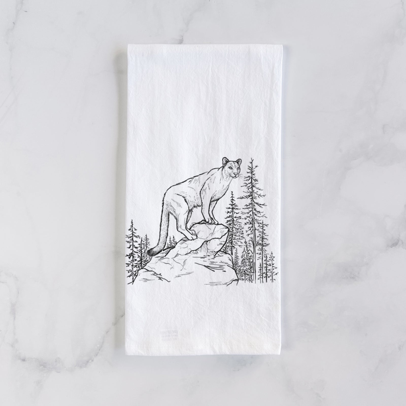 White cotton tea towel printed with a black ink illustration of a mountain lion standing on a rocky ledge among pine trees, on a marble counter beside a round wooden cutting board and greenery.