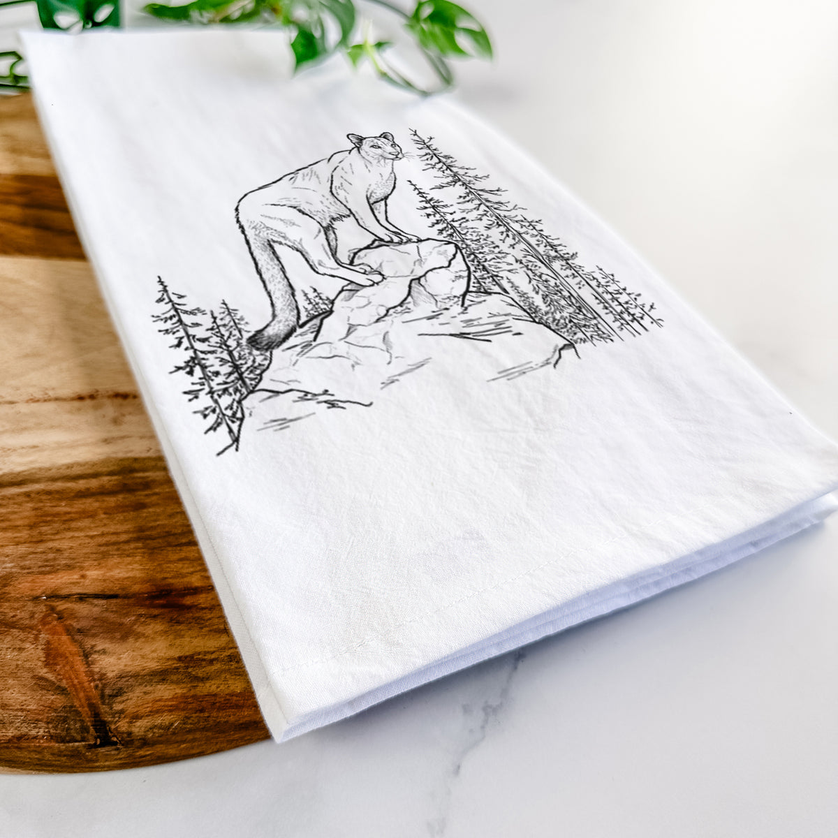 Close-up of a folded white tea towel on a wooden board, showing a black ink illustration of a mountain lion standing on a rocky ledge among pine trees.