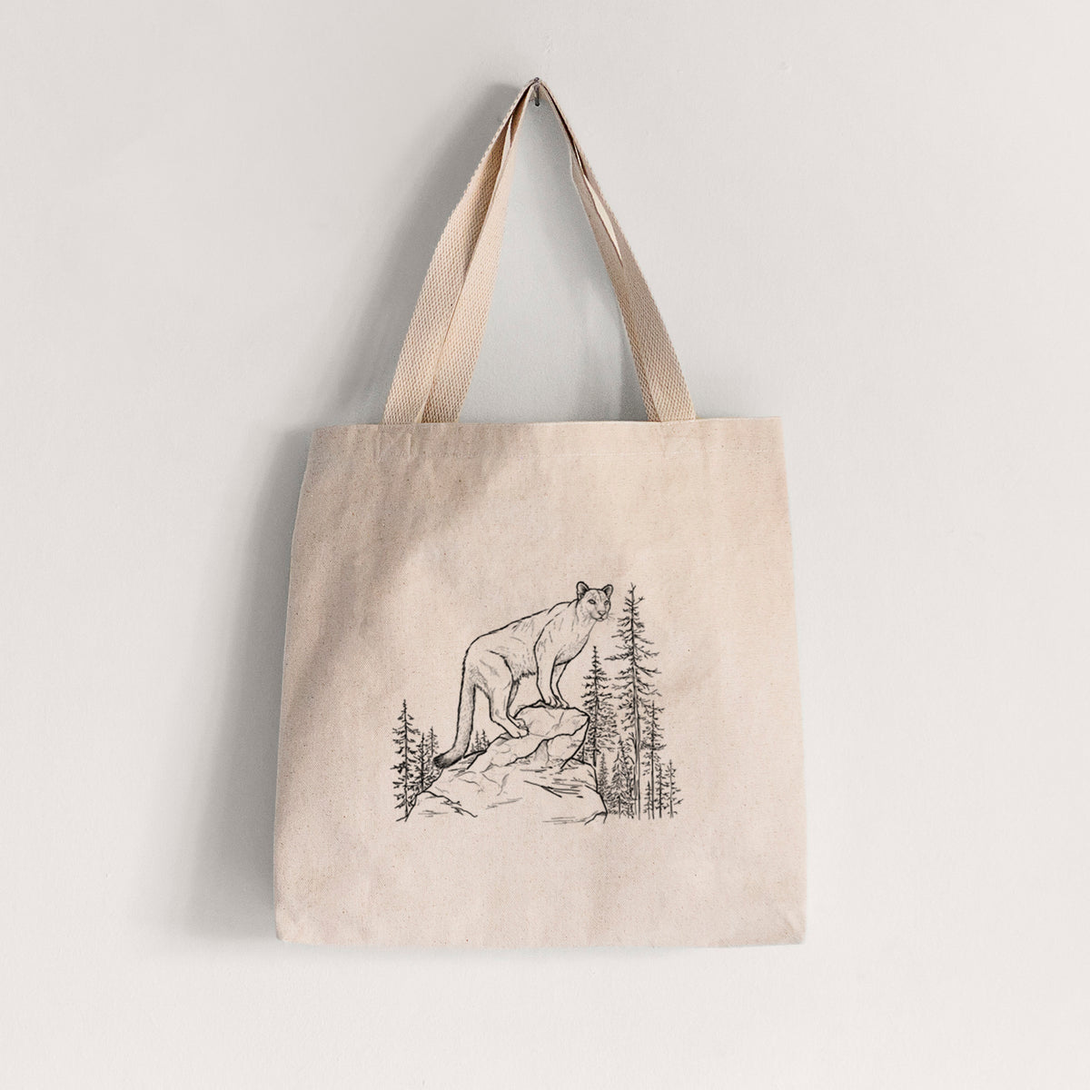 Natural canvas tote bag printed with a black ink illustration of a mountain lion standing on a rocky ledge among pine trees.