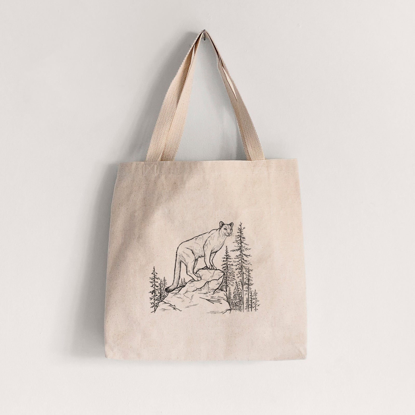Natural canvas tote bag printed with a black ink illustration of a mountain lion standing on a rocky ledge among pine trees.