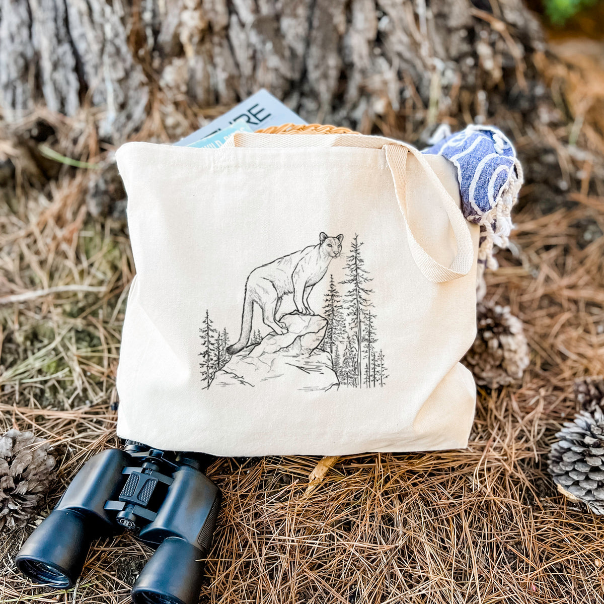 Canvas tote bag printed with a black ink illustration of a mountain lion standing on a rocky ledge among pine trees, resting on dry grass beside a pair of binoculars.